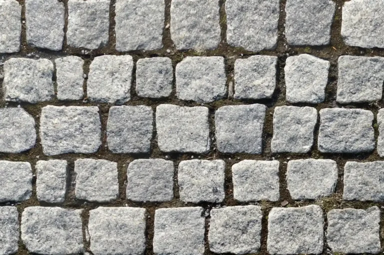 Close-up of a large stone pavement with light grey colored square shaped stones with earth between them