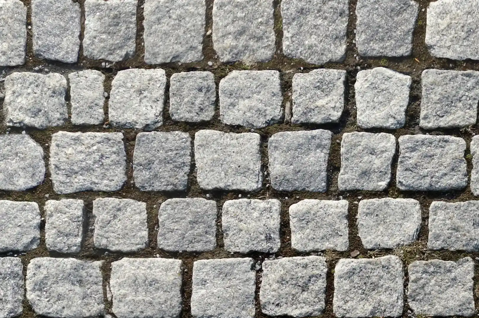 Close-up of a large stone pavement with light grey colored square shaped stones with earth between them
